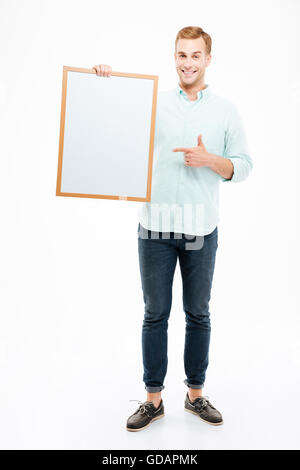 Cheerful young man holding blank white board and pointing on it over white background Stock Photo