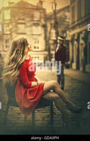 young beautiful blond woman sitting on chair in middle of cobble street Stock Photo