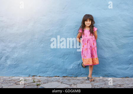 Full length portrait of pretty little girl looking at camera and smiling while standing against blue wall with copy space Stock Photo