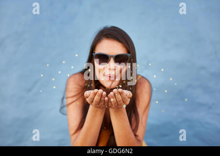 Close up shot of young woman blowing sparkles against blue background, focus on hands of female model. Stock Photo