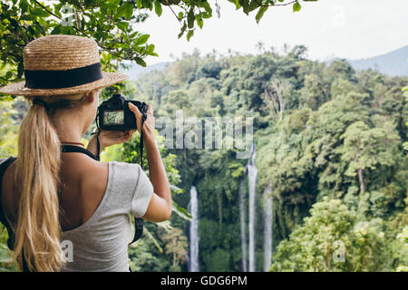 Rear view shot of female tourist photographing a waterfall with her digital camera. Young woman wearing hat taking a picture of Stock Photo