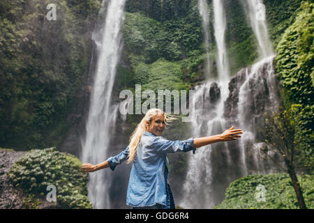 Rear view of happy young woman standing near waterfall with her hands raised. Female tourist enjoying by a water fall in forest. Stock Photo