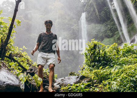 Outdoors shot of caucasian male hiker walking down the mountain trail. Young man hiking in forest with waterfall in background. Stock Photo