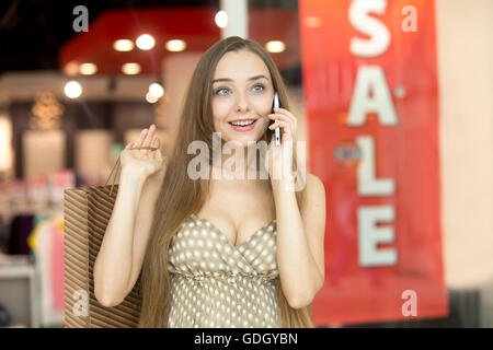 A portrait of a young beautiful woman standing in shopping centre talking on her mobile phone carrying shopping bag on her Stock Photo