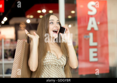 Candid portrait of young emotional joyful pretty woman in shopping centre talking on phone and shouting in surprise carrying Stock Photo