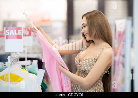 Profile portrait of joyful young pretty woman in shopping centre interior choosing clothes looking at cute pink summer apparel Stock Photo