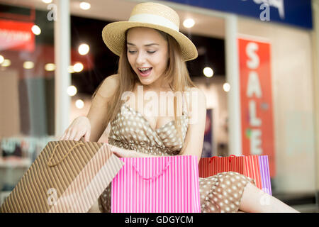 Excited young pretty woman in hat sitting in shopping centre with shopping bags. She is glad, looking at purchase Stock Photo