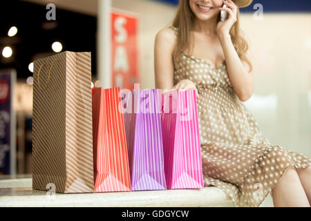 Close-up of beautiful smiling young shopper woman sitting with shopping paper bags talking on phone, making call. Focus on bag Stock Photo