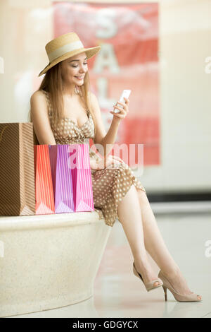 Beautiful happy woman wearing hat and dress sitting with legs crossed in shopping centre near clothing store with colorful Stock Photo