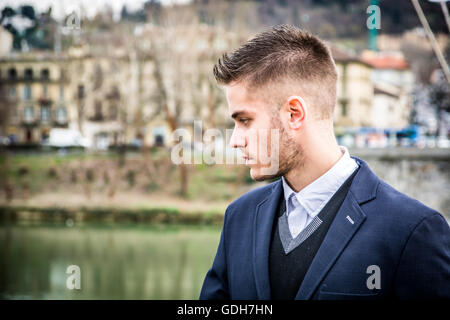 Three-quarter length of contemplative light brown haired young man wearing grey jacket and denim jeans standing beside picturesq Stock Photo