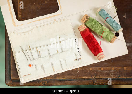 A basic sewing kit consisting of needles, a pin, and three spools of thread on a card laid on a wooden surface. Stock Photo
