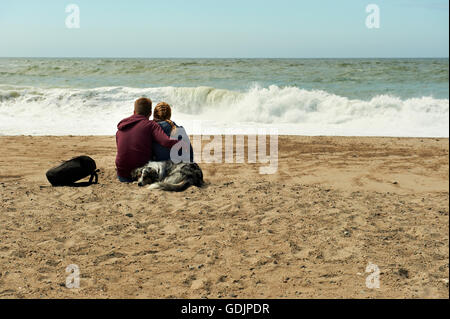 Young couple and their dog watching the waves breaking ont the beach Stock Photo