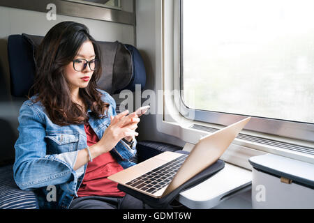 Cute Asian woman using smartphone and laptop on train, copy space on window, business travel or technology concept Stock Photo