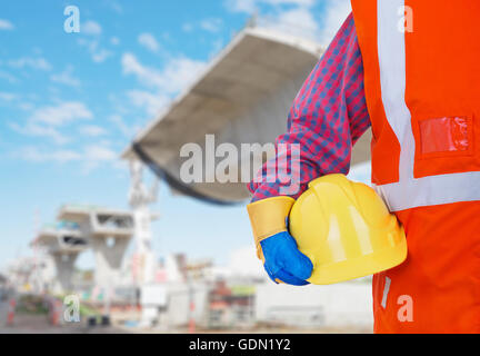 Safety Protective Work Equipment. Worker in orange vest holding yellow helmet against construction site as background Stock Photo