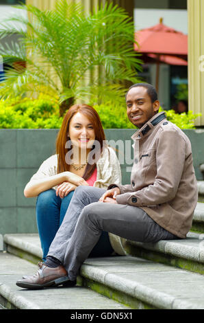 Interracial happy charming couple sitting on steps in front of building interacting and smiling for camera Stock Photo