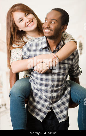 Interracial couple wearing casual clothes interacting having fun, man carrying woman on his back, both smiling happily, white studio background Stock Photo