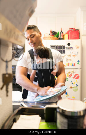 Father with baby in carrier, washing dishes Stock Photo