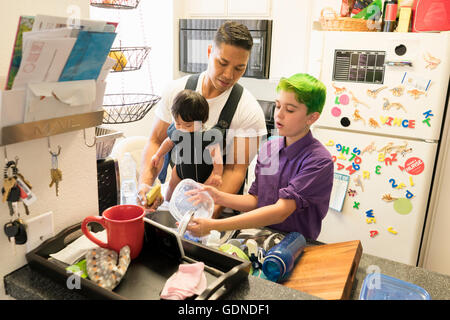 Son helping father with baby in carrier wash dishes Stock Photo