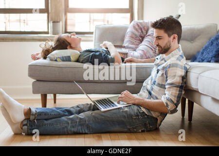 Young couple relaxing at home, young woman sleeping on sofa, young man using laptop Stock Photo