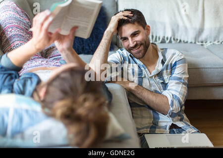 Young couple relaxing at home, young woman lying on sofa, young man sitting on floor, laughing Stock Photo