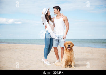Happy young couple hugging and smiling while walking with dog on the beach Stock Photo