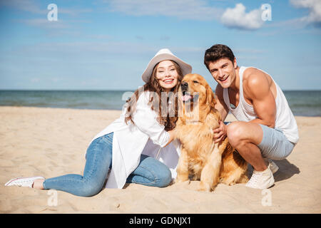 Happy young couple sitting and having fun with dog on the beach Stock Photo