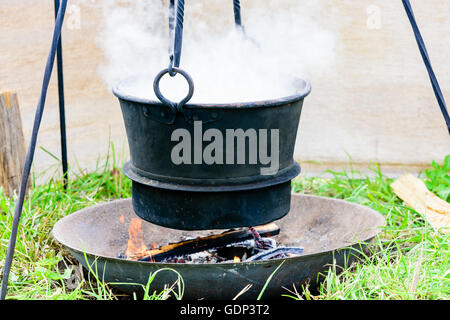 Boiling hot iron pot hanging over open fire. Water vapor coming up from the pot. Cooking or dying at a campsite. Stock Photo