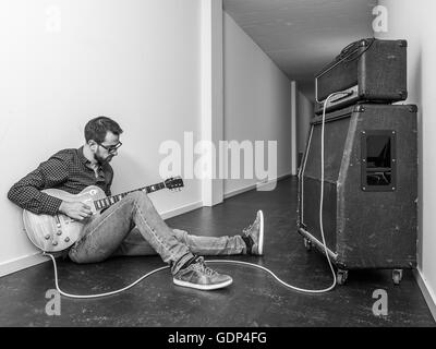 Photo of a man sitting playing his electric guitar in front of a large amplifier in a hallway. Black and white version. Stock Photo