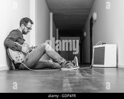 Photo of a man sitting playing his electric guitar in front of a small combo amplifier in a hallway. Black and white version. Stock Photo