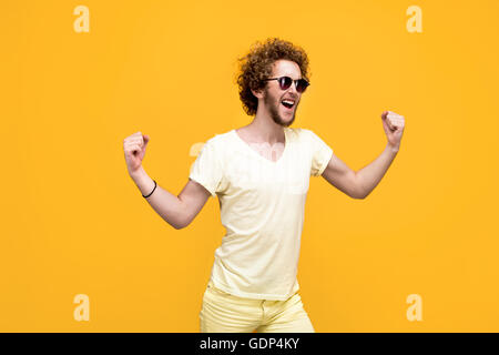 Portrait of curly-haired succeed young man in sunglasses and bermudas hands up. Isolate. Yellow background. Stock Photo
