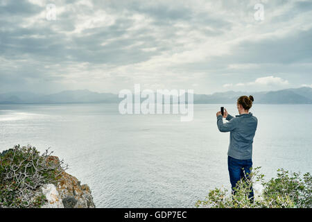Young man standing on cliff photographing with smartphone, Alcudia, Majorca, Spain Stock Photo