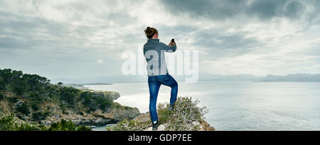 Panoramic view of young man standing on cliff photographing with smartphone, Alcudia, Majorca, Spain Stock Photo