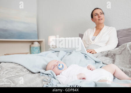 Mother sitting in bed using laptop while baby boy sleeps beside her Stock Photo