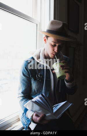 Young man at home, sitting on windowsill, drinking health drink, reading book Stock Photo