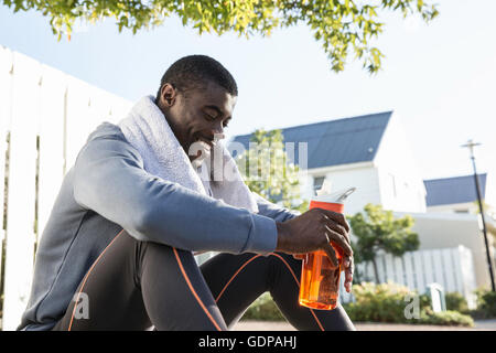 Man in residential area sitting on kerb holding water bottle looking down smiling Stock Photo