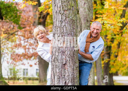 Couple, senior man and woman, flirting with each other playing hide and seek around a tree in fall tree Stock Photo