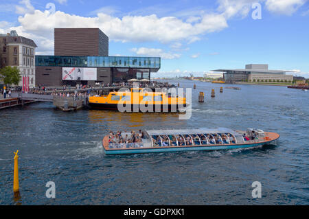 A canal cruise boat and a yellow harbour bus at the Royal Playhouse at Nyhavn in Copenhagen. The Royal Danish Opera House opposite further away. Stock Photo