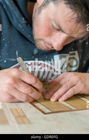A caucasian male in paint-splattered hoodie using a precision knife or blade and a stencil and working on a drafting table. Stock Photo