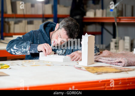 A man in paint-splattered dark hoodie busy scuplting a white plaster building model using a precision knife on a drafting table. Stock Photo
