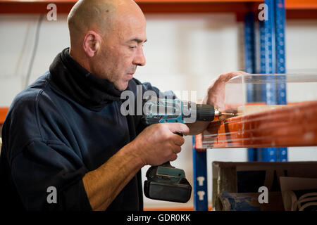 A man in a dark jacket drilling holes on one side of a transparent acrylic case using a cordless drill. Stock Photo