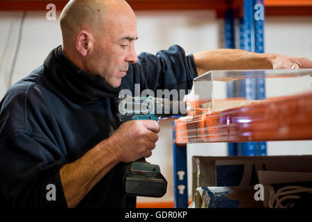 A man in a dark jacket drilling holes on one side of a transparent acrylic case using a cordless power drill. Stock Photo
