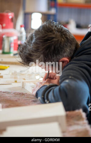 A man in a dark hoodie engraving a section of a white plaster building model using a precision knife on a drafting table. Stock Photo