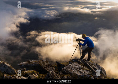 A landscape photographer setting up for his shot above the clouds on a steep cliff in the Scottish Highlands (Kintail) Stock Photo