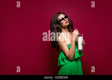 Portrait of beautiful young brunette in sunglasses and green dress with cocktail on rad background. Isolated Stock Photo