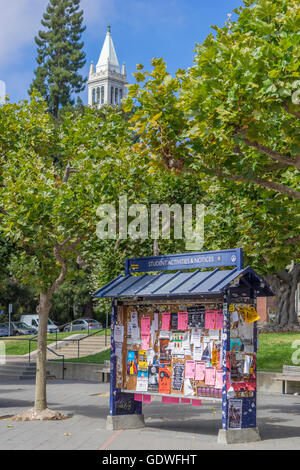 Many flyers posted on a bulletin board kiosk with campanile tower in background on Berkeley campus Stock Photo