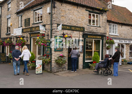 2 people pass while others buy ice creams at Hunters of Helmsley, an independent delicatessen - Helmsley, North Yorkshire, GB. Stock Photo