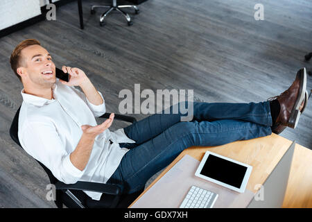 Top view of smiling confident young businessman sitting and talking on mobile phone at workplace Stock Photo