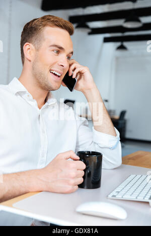 Cheerful young businessman drinking coffee and talking on mobile phone in office Stock Photo