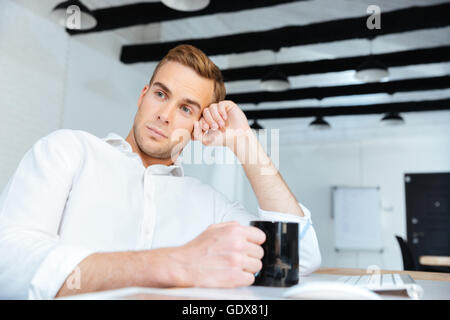 Thoughtful peaceful young businessman drinking coffee and thinking at workplace Stock Photo