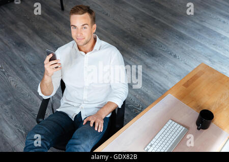 Top view of smiling relaxed young businessman sitting and using smartphone in office Stock Photo